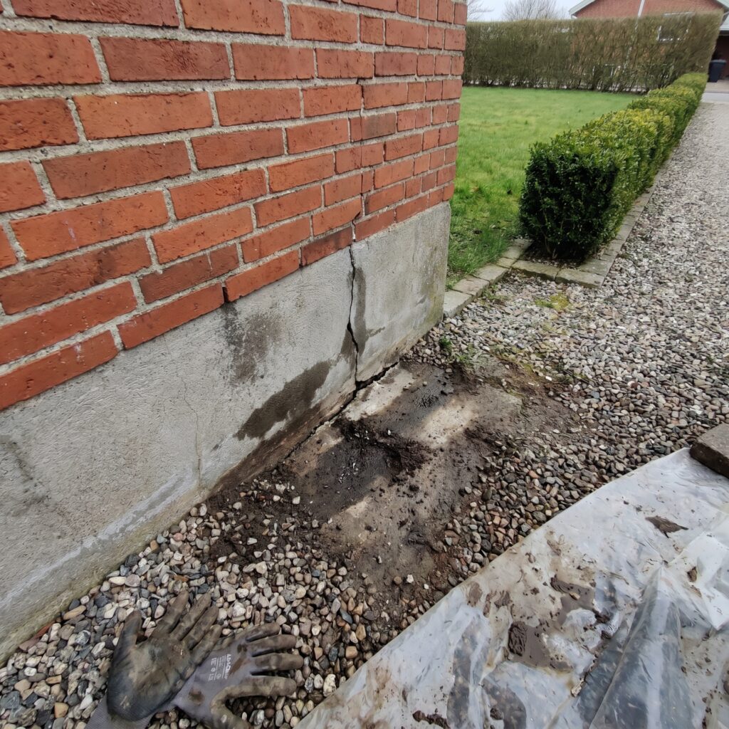 overlap-fundament, foundation_drain, detail, outdoor, fundament,sokkel,dræn,fugt,revner,kælder,omfangsdræn,beton,reparation, Nærbillede af sokkel med lille revne, fugtspor og grus langs et dansk hus., ved et parcelhus med græsplæne og lav hæk., dansk bolig, håndværk, renovering, vedligeholdelse, pris, tilbud, foundation_drain, detail, outdoor, fundament,sokkel,dræn,fugt,revner,kælder,omfangsdræn,beton,reparation
