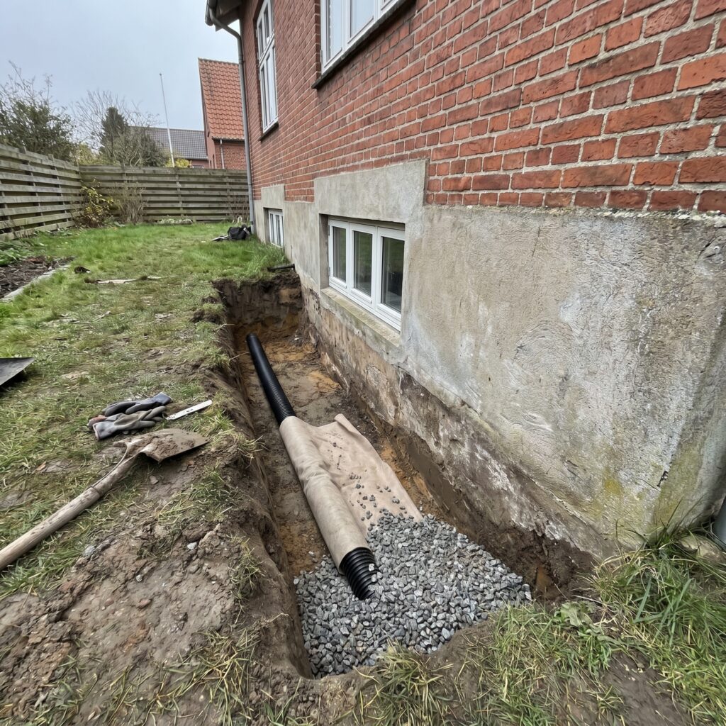 overlap-fundament, foundation_drain, detail, outdoor, fundament,sokkel,dræn,fugt,revner,kælder,omfangsdræn,beton,reparation, Detalje af drænrør med filterdug og singels langs fundament i åben rende., ved en ældre bolig med kælder., dansk bolig, håndværk, renovering, vedligeholdelse, pris, tilbud, foundation_drain, detail, outdoor, fundament,sokkel,dræn,fugt,revner,kælder,omfangsdræn,beton,reparation
