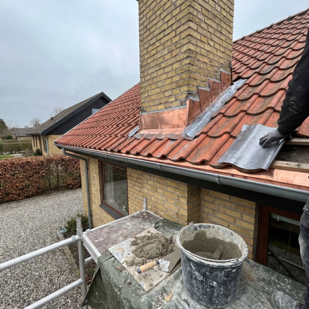 overlap-tag, roof_chimney, detail, outdoor, tag,skorsten,tagrender,inddækning,tagsten,rygning,vedligehold,reparation, Nærbillede af tagsten, inddækning og fuge omkring en muret skorsten., på et lavt parcelhus hvor arbejdet kan ses fra haven., dansk bolig, håndværk, renovering, vedligeholdelse, pris, tilbud, roof_chimney, detail, outdoor, tag,skorsten,tagrender,inddækning,tagsten,rygning,vedligehold,reparation