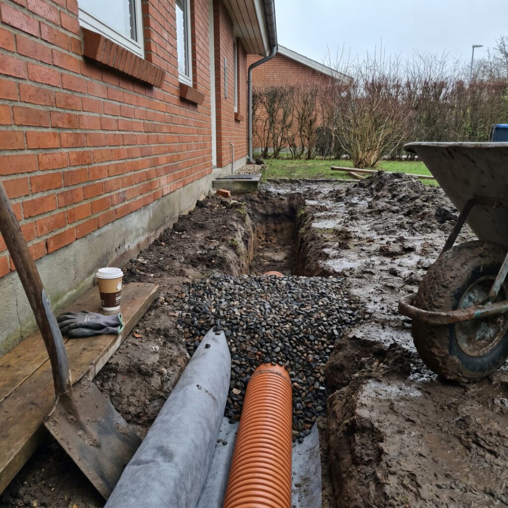 overlap-kloak-jord, sewer_groundwork, detail, outdoor, kloak,afløb,brønd,dræn,udgravning,jordarbejde,faskine,regnvand, Nærbillede af drænrør, geotekstil og singels langs hus., i gråvejr med realistisk våd jord., dansk bolig, håndværk, renovering, vedligeholdelse, pris, tilbud, sewer_groundwork, detail, outdoor, kloak,afløb,brønd,dræn,udgravning,jordarbejde,faskine,regnvand